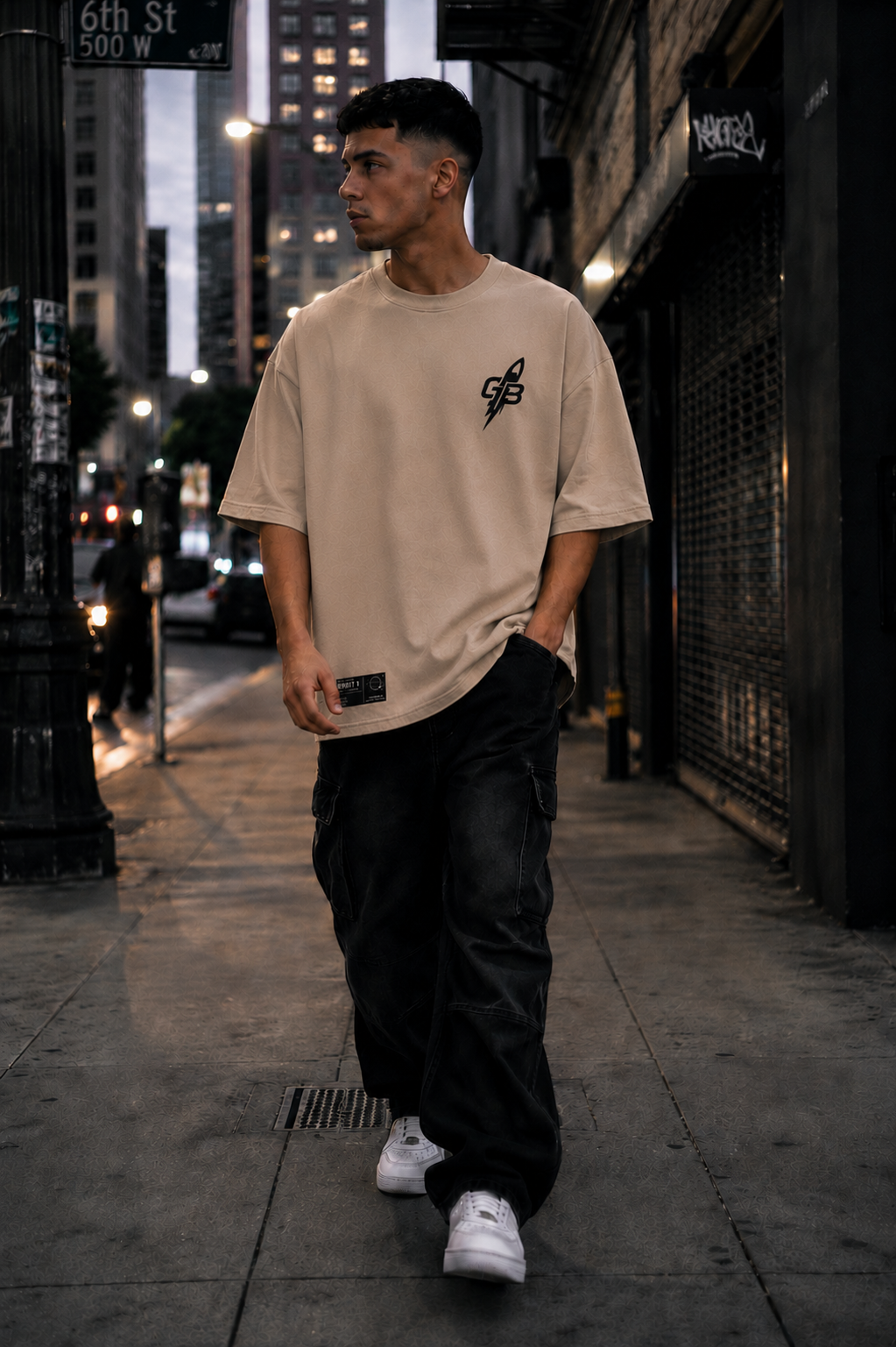 Male model walking through a city street wearing the GLXY Orbit 1 cream heavyweight oversized t-shirt featuring a black GB rocket logo and Orbit woven identifier.